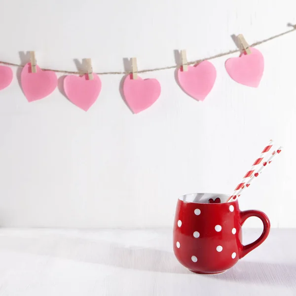 Красная чашка в белой точке на белом деревянном столе. Бумажные сердца висят на стене — стоковое фото