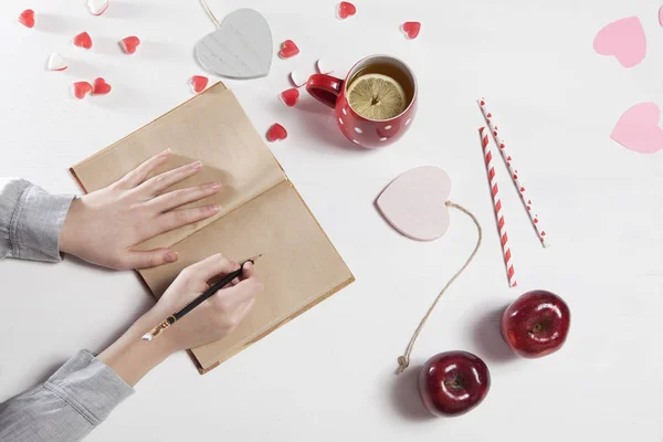 Manos de una mujer que va a dibujar con un lápiz en un cuaderno. En la mesa hay una taza de té con limón, manzanas y corazones de papel — Foto de Stock