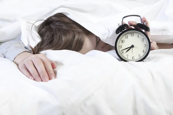 Girl, wrapped in a white blanket, puts out her hand to turn off the alarm. There is eight hours on the alarm clock. — Stock Photo, Image