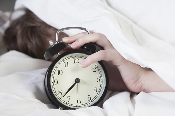 Girl, wrapped in a white blanket, puts out her hand to turn off the alarm. There is eight hours on the alarm clock. — Stock Photo, Image