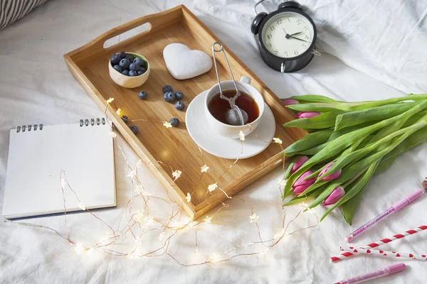 Bricka med te och pepparkakor i form av hjärta och en bukett tulpaner på sängen — Stockfoto