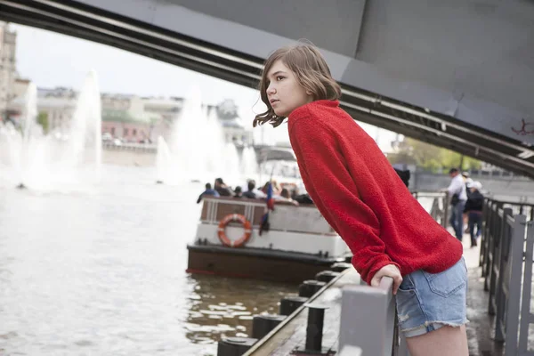 Uma jovem com uma camisola de lã vermelha e calções de ganga. Ela encostou-se ao corrimão ao lado do canal. — Fotografia de Stock