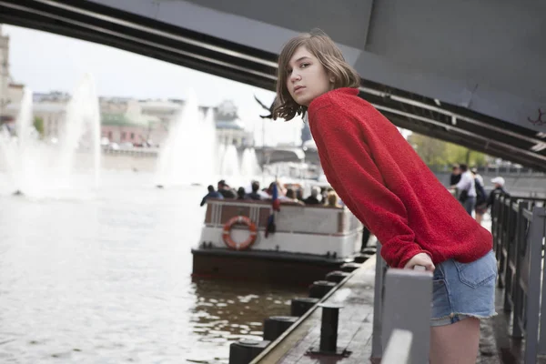一个穿着红色羊毛毛衣和牛仔裤短裤的年轻女孩。她靠在运河旁的栏杆上 — 图库照片