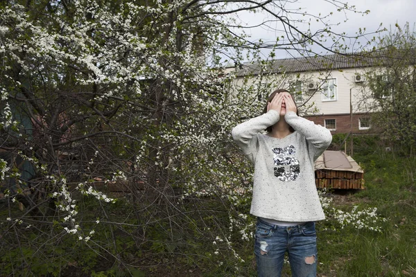 Meisje in een witte trui sluit dromerig haar ogen uit de felle zon in de tuin in de buurt van de bloeiende kersen. — Stockfoto