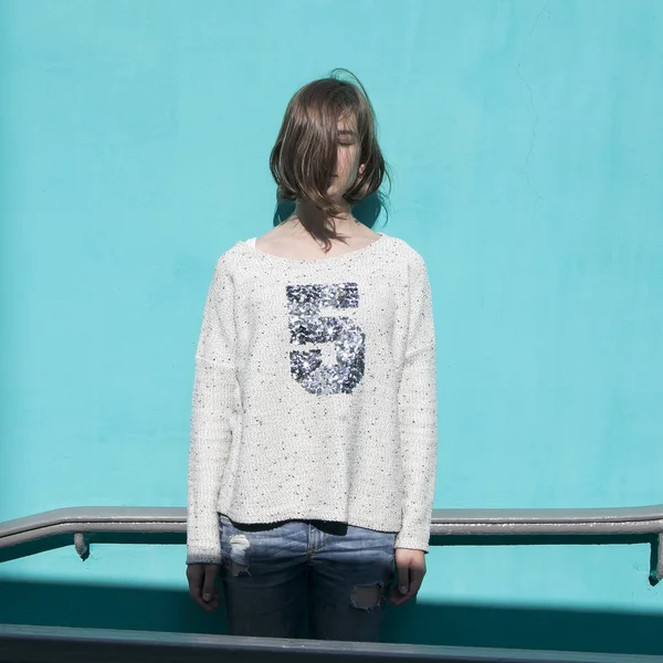 Girl in a white sweater dreamily closes her eyes from the bright sun opposite the blue wall in the underpass — Stock Photo, Image