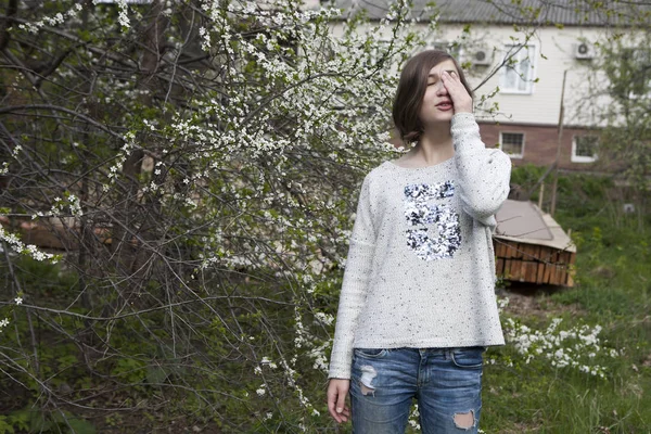白いセーターの女の子はうっとりと咲く桜の近くの庭の明るい太陽から目を閉じる. — ストック写真