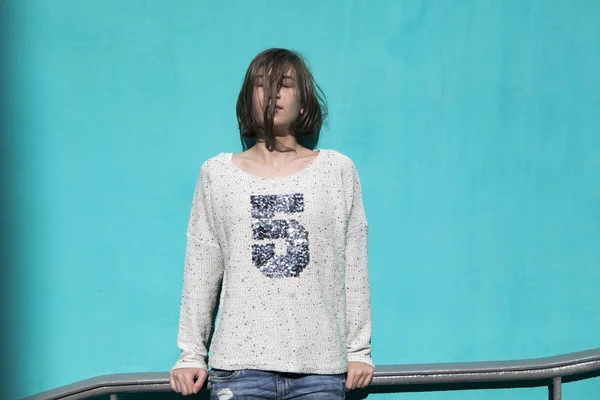 Girl in a white sweater dreamily closes her eyes from the bright sun opposite the blue wall in the underpass — Stock Photo, Image