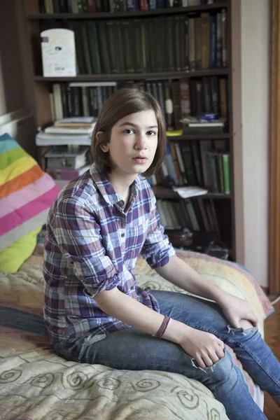 The A thoughtful girl in a plaid shirt is sitting on the bed in front of a bookcase — Stock Photo, Image