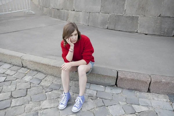 Jong meisje in een rode wollen trui en jeans broek zit op de stappen — Stockfoto