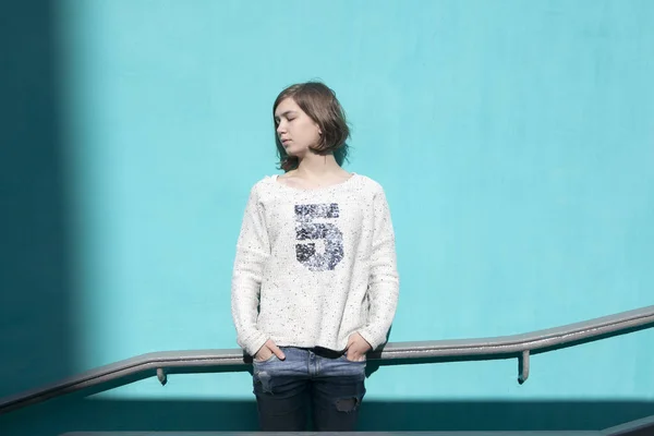 Girl in a white sweater dreamily closes her eyes from the bright sun opposite the blue wall in the underpass — Stock Photo, Image