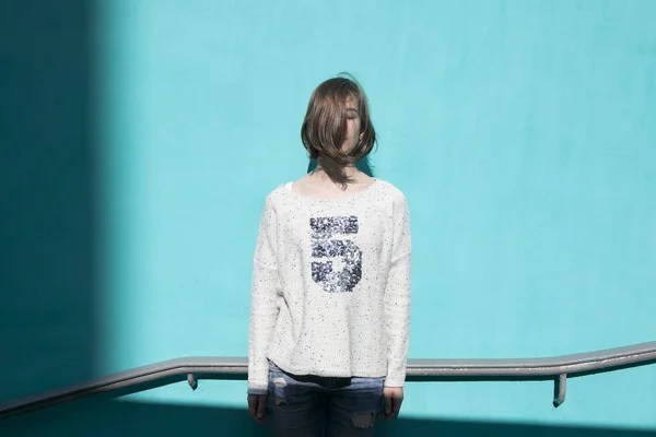 Girl in a white sweater dreamily closes her eyes from the bright sun opposite the blue wall in the underpass — Stock Photo, Image