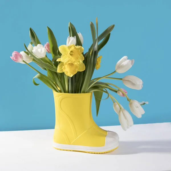 The Yellow rubber boots with a bouquet of flowers of yellow daffodils and white and pink tulips. Garden accessories. — Stock Photo, Image