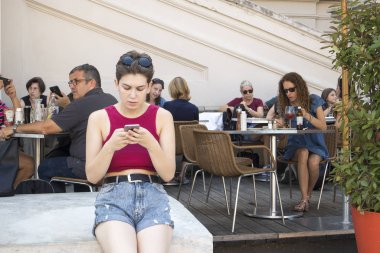 Şehir merkezinde sonbahar caddeleri kalabalıktı. İnsanlar bir sokak kafesinde yemek yiyor. Kız cep telefonuna bakıyor.