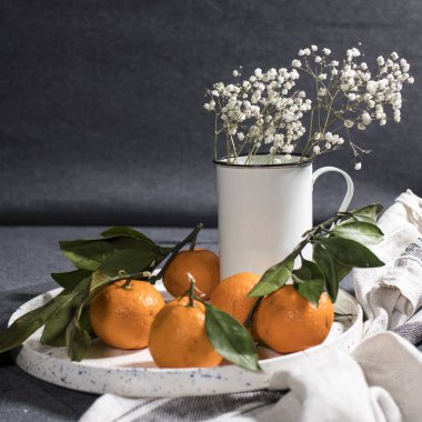 Beyaz seramik tabakta yeşil yapraklı mandalinalar ve gri mutfak havlusu.