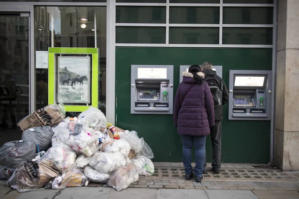 People withdraw cash from an ATM on the street Strand. Nearby is a huge pile of garbage. — ストック写真
