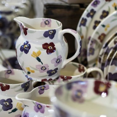 Mağazadaki bir rafta beyaz seramik tabaklar. Desenci seramik plakalar. Masa takımı ve yeme konsepti. Masa için güllü beyaz sürahi şarap için bardaklar ve su için bir şişe.