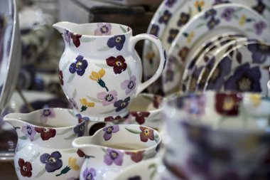 Mağazadaki bir rafta beyaz seramik tabaklar. Desenci seramik plakalar. Masa takımı ve yeme konsepti. Masa için güllü beyaz sürahi şarap için bardaklar ve su için bir şişe.