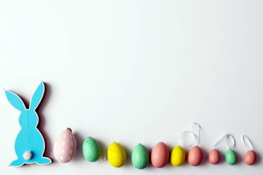 Beyaz arka planda, üzerinde plastik yumurtalar olan mavi, düz bir Paskalya tavşanı yatıyor. Boşluğu kopyala