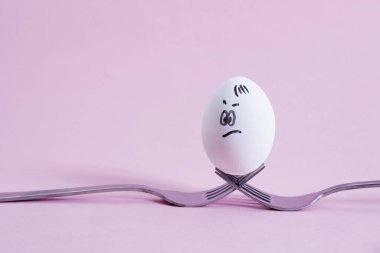 Çapraz çatalların üzerinde beyaz bir yumurta, pembe arka planda bir yumurta standı gibi duruyor. Uzayı kopyala Kavram