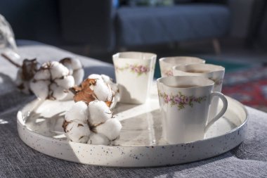 Pamuk çiçekli beyaz tabure ve porselen çay fincanlarıyla dolu tepsi. Arka planda pencere gölgesi olan bir tabure..