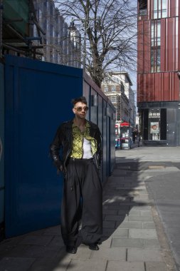 Londra, İngiltere - 15 Ocak 2020, güneş gözlüklü esmer adam, siyah geniş pantolon ve beyaz tişörtlü mavi bir duvara poz veren sarı tişörtlü