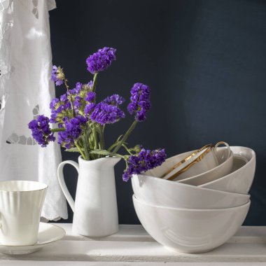 Bir buket limonyum ve beyaz tabaklar: porselen süt sürahisi, kahve fincanı, koyu mavi arkaplanda beyaz bir rafta kaseler. Mutfak.
