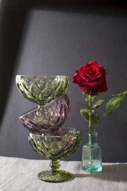 Koyu yeşil ve mor camlı krema için üç vazo siyah arka planda gölgeli bir şekilde üst üste dururlar. Vazoda koyu kırmızı bir gül var.