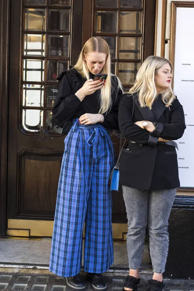 ロンドン イギリス 2020年1月15日路上でファッショナブルな人々 ストリートスタイル 二人のブロンドの女の子が友達を待っている 青いチェックパンツの男が電話を見て — ストック写真