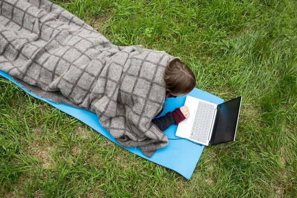 一位身着灰色羊毛衫的女孩 躺在河边草地上的一块运动垫上 一边用笔记本电脑干活 — 图库照片