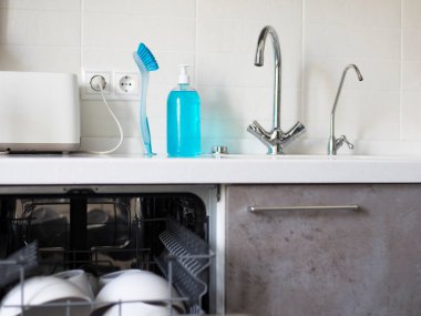 Mutfaktan bir parça. Entegre lavabosu olan yapay taş tezgah. İki musluk. Bir tane filtrelenmiş su için. Elleri ve bulaşıkları yıkamak için sıvı sabun. Açık bulaşık makinası. Beyaz kiremitli duvar.