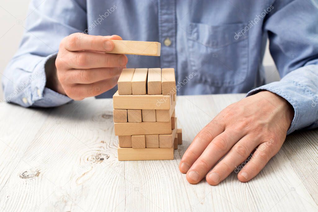 Men's hands in a blue office shirt are folding a pyramid of wooden bars, which can be inscribed. Fragment. Unrecognizable person. Against the background of a beige table with a wooden texture.