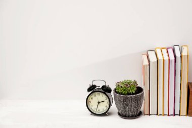 Uzayı kopyala Aksesuarları ve beyaz masası olan bir işyeri çalışanı. Çalar saat, seramik bir tencerede sulu bir saat ve bir öğrencinin masasında bir yığın kitap..