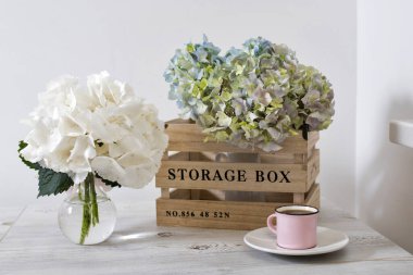 Depodaki ahşap kutuda yeşil ve mavi pastel ortancayla büyük bir dal ve yuvarlak cam vazoda beyaz ortanca ve bej bir masada kahve bulunan küçük pembe kupa..