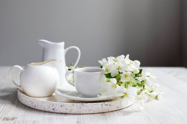 Elma ağacının çiçek açan bir dalına sahip vazo. Beyaz tabak seti: bir fincan kahve, süt sürahisi, bir sürahi, masanın üzerinde beyaz bir duvarın arka planında duran bir tepsi. Boşluğu kopyala