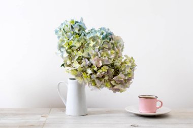 Bej bir masada beyaz bir sürahi içinde yeşil ve mavi pastel ortancayla büyük bir dal. Beyaz bir sürahide yeşil ve mavi pastel ortancalı büyük bir dal bej bir masada bir fincan kahve ve süt. Boşluğu kopyala
