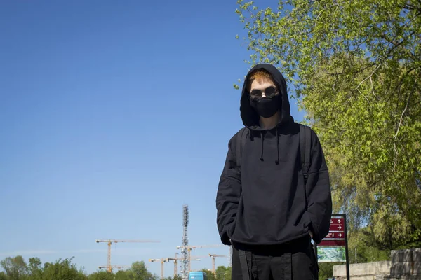 Young man with orange dyed hair in a black hoodie with trousers and sports boots in a protective mask and black sunglasses against a blue sky. Urban view. Quarantine street style.