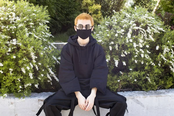 Young man with orange dyed hair in a black hoodie with trousers and sports boots in a protective mask and black sunglasses against a blue sky. Urban view. Quarantine street style.