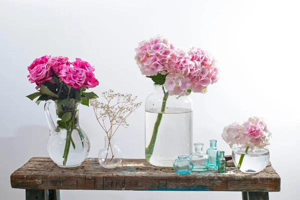 A bouquet of red roses in a tin bucket, multicolored pastel colored hydrangeas in a glass jar, gypsophila in a flask and a set of small bottles on the old surface of a wooden bench