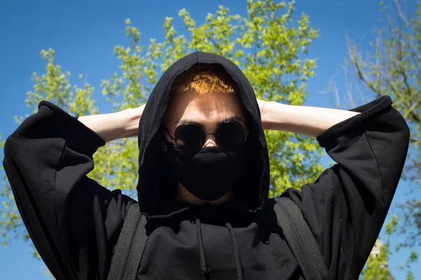 Young man with orange dyed hair in a black hoodie with trousers and sports boots in a protective mask and black sunglasses against a blue sky. Urban view. Quarantine street style.