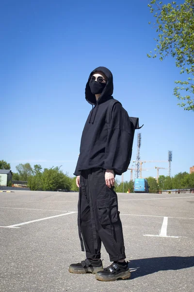 Young man with orange dyed hair in a black hoodie with trousers and sports boots in a protective mask and black sunglasses against a blue sky. Urban view. Quarantine street style.