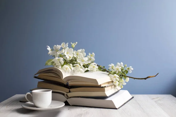 A branch of a blossoming apple tree or cherry on an open book and cup of coffee on a background of a blue wall. Concept - online learning. Copy space. Education on-line