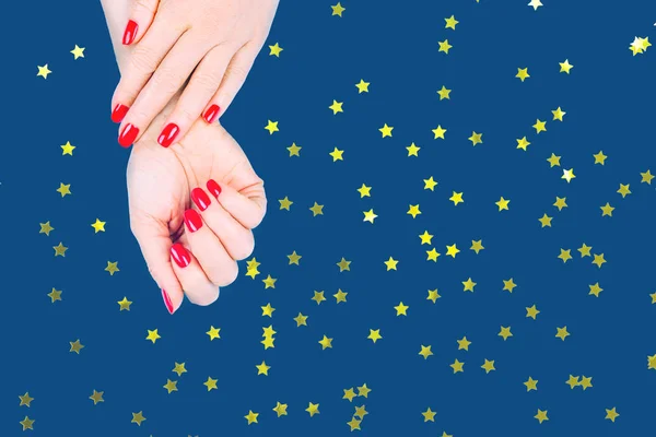 Mains de femme avec manucure rouge élégant sur fond bleu. Concept de vacances, fête et beauté avec confettis en forme d'étoile dorée festive — Photo