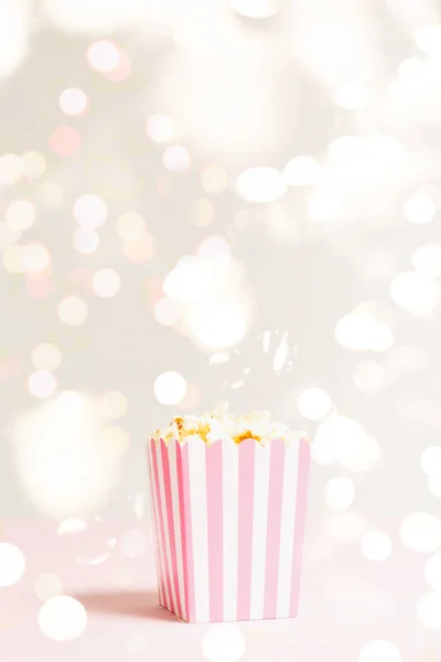 爆米花包，白色和粉红色条纹在节日灯火管制的背景。 空荡荡的简约背景 浪漫电影之夜和小吃的概念 — 图库照片