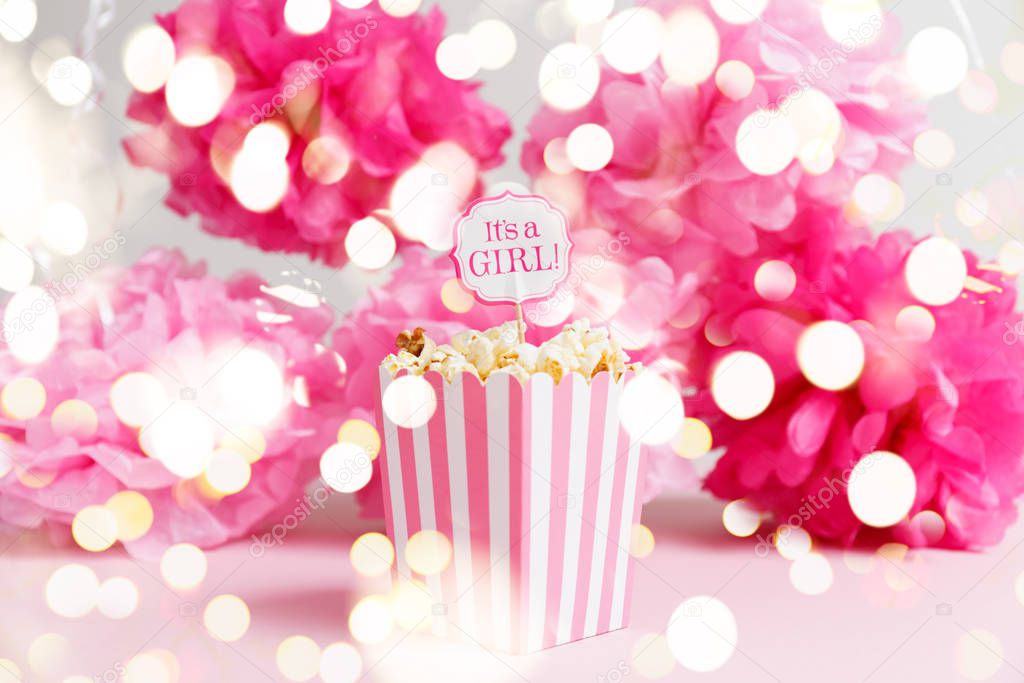 Its a girl sign in a popcorn bag at the baby shower party. Paper flowers background. Baby shower celebration concept with festive holiday bokeh