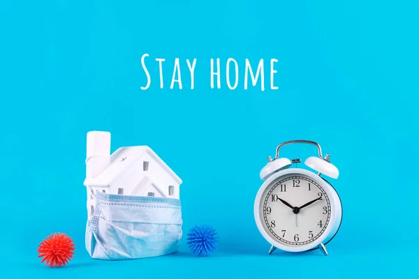 Cute little house covered with medicine mask, clock and plastic balls aka viruses on the blue background with Stay home wording. Epidemic, social isolation, coronavirus COVID-19 concept