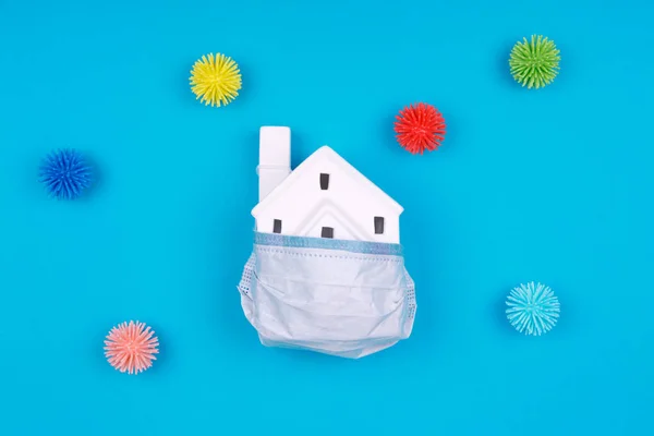 Cute little house covered with medicine mask and plastic balls as viruses on the blue background. Epidemic, social isolation, coronavirus COVID-19 concept