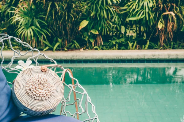 Trendy round crossbody straw bag with seashells and flower on the beautiful lounge chair near a pool