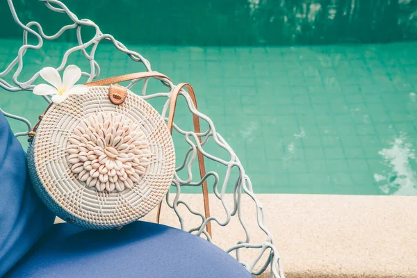 Trendy round crossbody straw bag with seashells and flower on the beautiful lounge chair near a pool