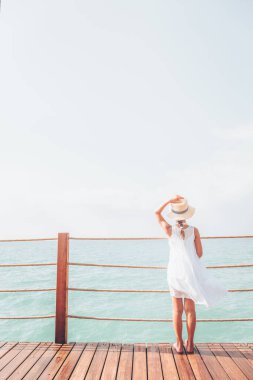 Beyaz elbiseli bronzlaşmış kız ahşap iskelede deniz manzarasının tadını çıkarıyor.