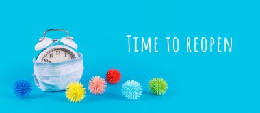 Tıbbi maskeli çalar saat ve mavi arka plandaki virüsler gibi plastik toplar. Coronavirus salgınının azaltılması, yeni normal ekonomiye geçiş, ekonominin yeniden açılması konsepti